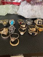 Lot of 13 mixed rings laid out on a black surface with crystal quartz in background, showing various styles, colors, and stones, including turquoise, pearl, clear stones, black enamel, and citrine-like stones. Two front rings missing stones.