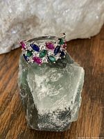 Photo of silver multi-coloured gemstone ring on a greenish crystal showing marquise blue, pink, green stones and clear small stones on band.