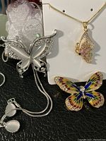 Photo showing all three jewelry pieces: gold-tone butterfly necklace with 22 cm chain, silver-tone butterfly brooch with white accents, and colorful enamel butterfly brooch