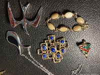 Photo showing all five jewelry items on black textured background: silver-tone necklace with teardrop pendant, gold-tone brooch with blue stones, enamel Christmas tree brooch, beige stone bracelet, and silver abstract pendant.