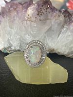 Front view of the silver ring showing oval moonstone surrounded by cubic zirconia stones, placed on a greenish stone with purple crystals in the background.