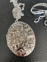 Close-up of oval silver-tone locket necklace with intricate etched scene and attached chain on black background