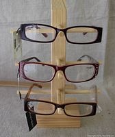 Front view of three pairs of reading glasses on wooden stand showing purple, red with rhinestones and brown frames with tags attached.