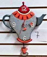 Full front view of the teapot clock showing the body as a grey teapot with red and pink details and black and white clock hands ending in small pink teacups.
