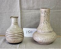 Two ceramic items side by side on a fabric surface: a ridged and speckled carafe with spout, and a taller vase with a wide base and narrow neck with cracked texture.