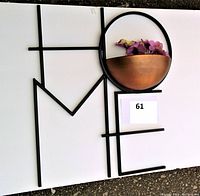 Metal wall art spelling 'HOME' with black painted letters and a copper-tone half-sphere planter as the letter O containing purple flowers (not included). The planter is affixed within the circular frame forming the O.