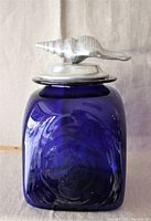 Front view of deep cobalt blue glass canister with detailed cast aluminum seashell stopper on top.