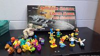 Photo of the Battlestar Galactica board game box, plush teddy bear, various Simpsons figurines and assorted animal figurines displayed on a table.