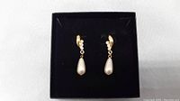 Pair of gold-tone drop earrings with pearl teardrop and rhinestones displayed in a black box against white background.