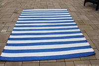 Blue and white striped plastic patio rug laid flat on outdoor stone surface, showing full length and pattern.