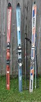 Three pairs of alpine skis (two Dynastar and one Kastle) and black ski poles arranged vertically against a wooden fence.