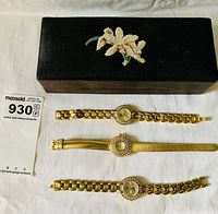 Top view of black needlepoint floral design box and three gold-tone ladies watches laid out horizontally below it showing different strap types and crystal accents around watch faces.