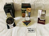 Three men's watches displayed in front of their boxes: George stainless steel watch with black dial, Timex Indiglo watch with white dial and expansion band, and Quartz calculator watch with red strap.