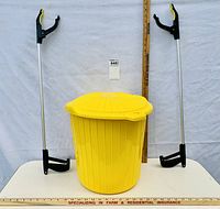 Front view of two pickup and reaching tools standing on either side of a yellow plastic garbage can with latching lid.