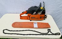 Full view of Husqvarna chainsaw body with attached rear handle, front handle and black top cover. Orange bar cover with Husqvarna branding and separate saw chain laid out in front.
