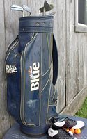 Blue Labatt's Blue branded golf bag with visible wear marks, standing upright outdoors, accompanied by golf shoes and golf balls on the ground.