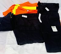 Overall and work pants laid on a white surface showing the orange overall with yellow reflective stripes and four pairs of black work pants with tags