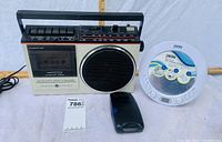 Three vintage radios shown side by side on white fabric background with measurement tape behind them. General Electric cassette radio/player on left, Otomus portable radio in middle front, JWIN alarm clock radio on right.