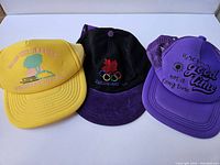 Photo showing three vintage hats including a yellow Camp Inn Resort hat, black Olympics hat with maple leaf and rings, and a purple slogan hat