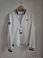 Front view of a white Wimbledon jacket with tags, showing full zipper and logo patches on chest and sleeve.