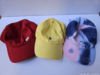 Photo showing three hats side by side: red Carhartt, yellow Polo Ralph Lauren, and blue/pink KITH tie-dye.