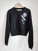 Front view of black Off-White sweatshirt showing blue daisy flower and branded tape detail on chest.