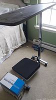 Full view of the metal adjustable bedside table with rectangular black top, mounted on a single metal stand with wheels and a black square cushion on base.