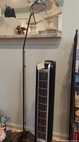 Full view of bendable floor lamp standing next to Lasko tower heater fan against a wall.