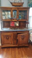 Full view of wooden two-piece server cabinet with white marble top on lower section, upper section with glass doors and mirrored back, decorative carvings, placed on wooden floor.
