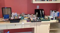Photo of desk surface showing assorted office supplies including pens, pencils, scissors, stapler, printer paper, ink cartridges, tape, and other small items.