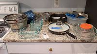 Overview of kitchen items on countertop including colanders, glass baking dishes, pans, springform pan, plastic container and apron.