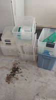 Group of clear and gray plastic stackable storage totes arranged on a concrete floor, some lids on, some placed to the side, stacked in two piles.
