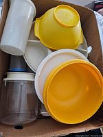 Photo showing a box with assorted vintage Tupperware including large yellow and white mixing bowls, a clear plastic container with lid, and other bowls.