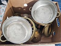 Box containing two round metal cooking pots with handles and several brass items including lids and smaller pieces.