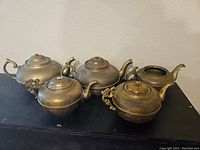 Five vintage brass teapots arranged on black surface showing various sizes and handle/spout designs.