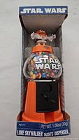 Full front view of the M&M dispenser in box showing orange base, clear globe with Star Wars logo and colorful M&M candies, topped with orange M&M Luke Skywalker figure.