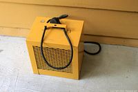 Yellow rectangular garage style heater with a black carrying handle and power cord placed on a concrete floor against a yellow wall.