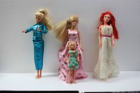 Four dolls shown standing side by side: Barbie in turquoise pantsuit, Midge in pink gown, smaller Francie in patchwork dress, and Skipper in white gown with pink belt.