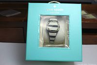 Kate Spade New York watch visible through turquoise box window, black and white striped band, gold-tone round bezel.