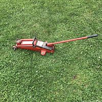 Red hydraulic trolley jack lying on grass, showing full profile with handle extended.