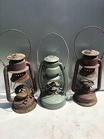 Front view of three vintage lanterns: two red rusty Beacon SMP lanterns flanking a green Embury Air Pilot lantern. All missing glass globes and handles intact.