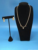 Gold tone necklace displayed on black jewelry bust with gold tone pierced earrings displayed on a stand beside it. Necklace features clear rhinestones with a dangling square pendant.