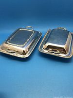 Two rectangular silver plated lidded serving dishes side by side showing general shape and style with lid handle and tray handles visible.
