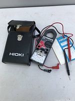 Hioki branded black protective carrying case open showing the clamp meter, test leads, and instruction manual on a white surface