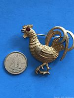 Gold-tone rooster brooch shown alongside a Canadian dime for size reference. The brooch features a red stone eye and detailed textured feathers with curled tail feathers.