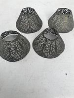 Four silver-tone vintage pierced metal lamp shades with floral and scroll cutout design, arranged on white surface