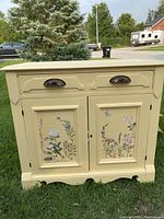 Front view of antique washstand showing cream paint and floral decoupage on cabinet doors with two drawers on top and metal handles.