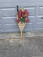 Photo of a vintage wooden snowshoe leaning against a grey garage door, decorated with pine sprigs, pinecones, and a large red and black checkered bow for Christmas.