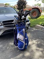 Blue golf bag standing on pavement with set of right-handed clubs inserted, 4 headcovers on larger clubs