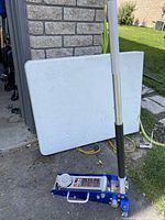 Blue Big Red 1.5 ton hydraulic service jack with foam grip handle leaning against a white table or board outside near a brick wall and grass.